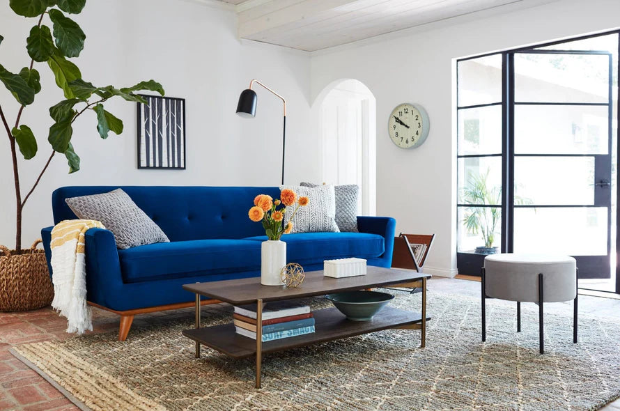 A modern living room featuring the Carson Sofa in blue, adorned with several patterned pillows and a cozy throw blanket. A wooden coffee table, embellished with flowers and books, rests on a textured rug. In the background, a large plant and artwork add charm, while a clock adorns the wall near a glass door.