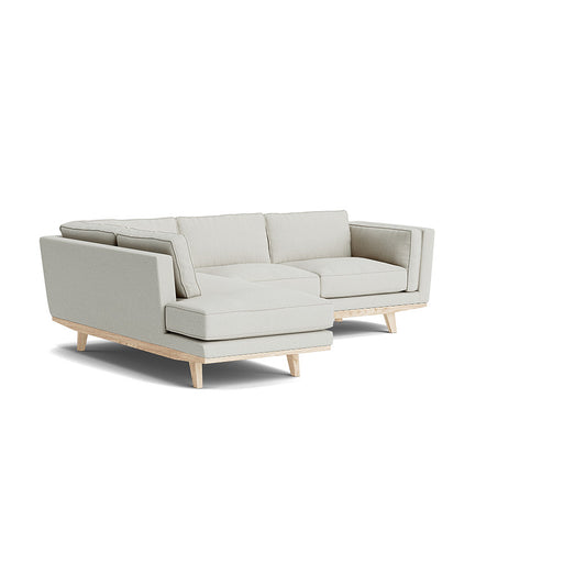 The Kirnik Bumper Sectional in Natural Latex is a light gray L-shaped sofa with wooden legs, set against a plain white backdrop. It boasts a modern design with plush cushions and clean lines.