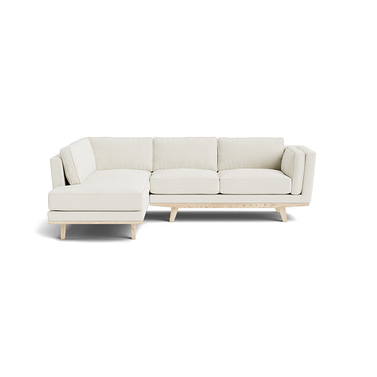 The Kirnik Bumper Sectional in Natural Latex is a modern sectional sofa featuring clean lines and wooden legs. In light beige, this sofa includes a chaise on the left side, offering ample seating space and a minimalist design, all set against a plain white background.