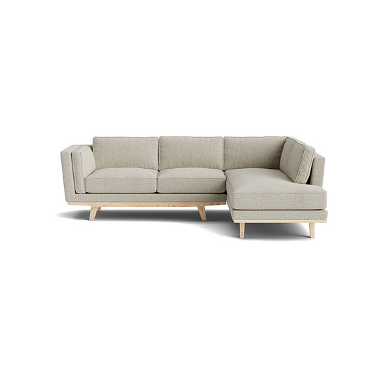 Introducing the Kirnik Bumper Sectional in Natural Latex, an L-shaped sectional sofa on a white background with wooden legs. This piece boasts clean lines and a modern design, complete with plush cushions on both the seats and backrests in a light gray hue.