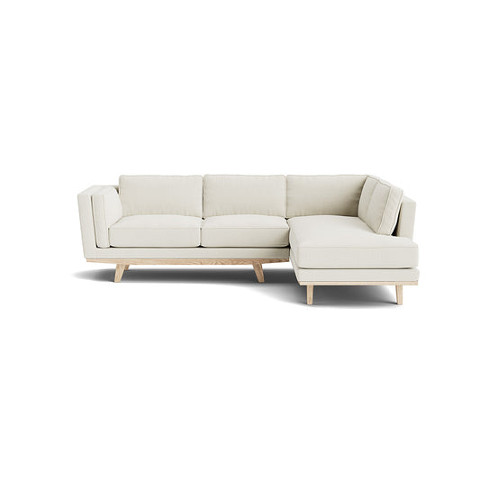 The Kirnik Bumper Sectional in Natural Latex is a cream-colored, L-shaped sofa with a wooden base and legs, set against a plain white background. It features a modern design with clean lines and plush cushions.