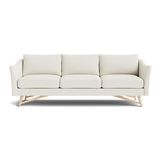 The Gio Sofa in Natural Latex is a modern cream-colored piece, fitted with three cushions and wooden legs. It features angled arms and a minimalist design, all set against a white background.