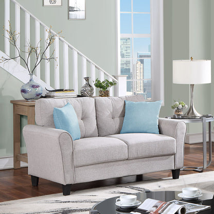 A cozy living room showcases the Modern Living Room Loveseat Linen Upholstered Couch in light gray with blue pillows. A wooden console table with vases and plants sits by the staircase, a window reveals a cityscape view, and a round side table with a lamp alongside a rug completes the space.