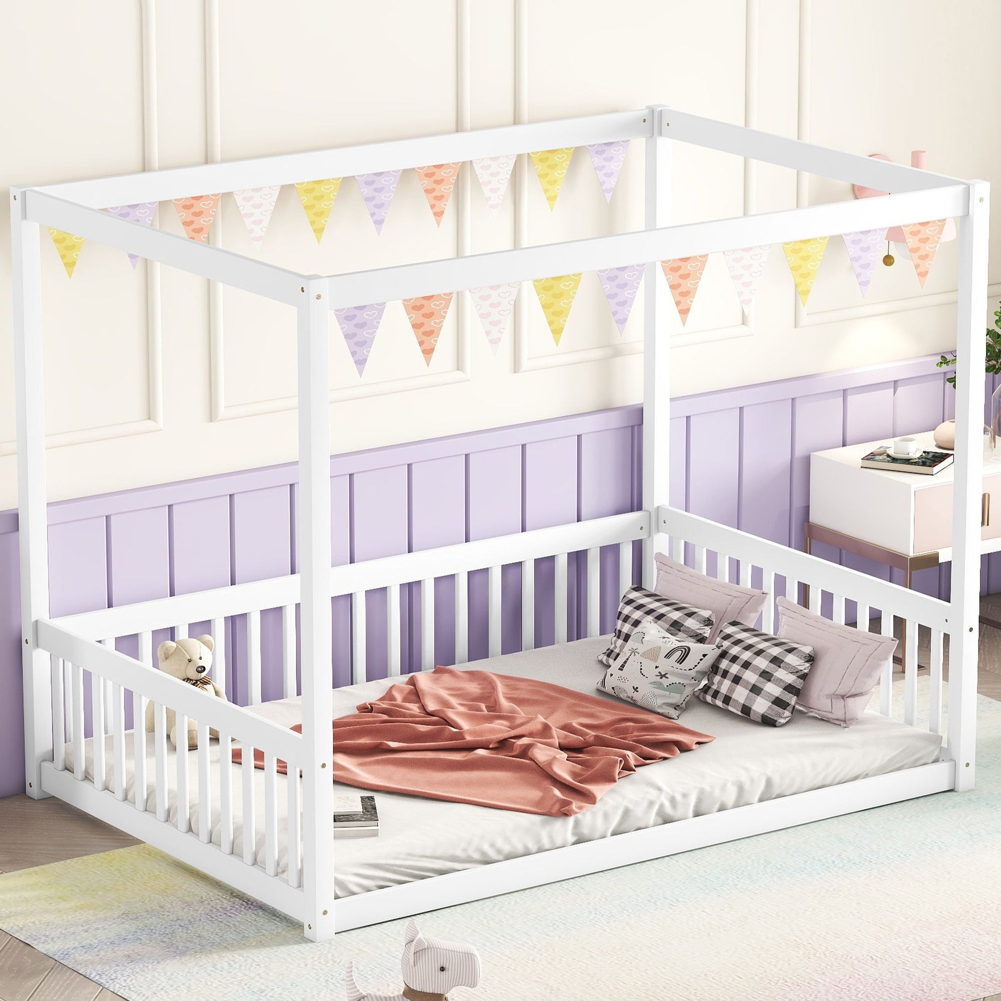 A Canopy Frame Floor Bed with a fence and guardrails in white stands against a purple wainscoted wall, adorned with pink and gray bedding. Above it hangs pastel triangular bunting, while nearby rests a plush toy.