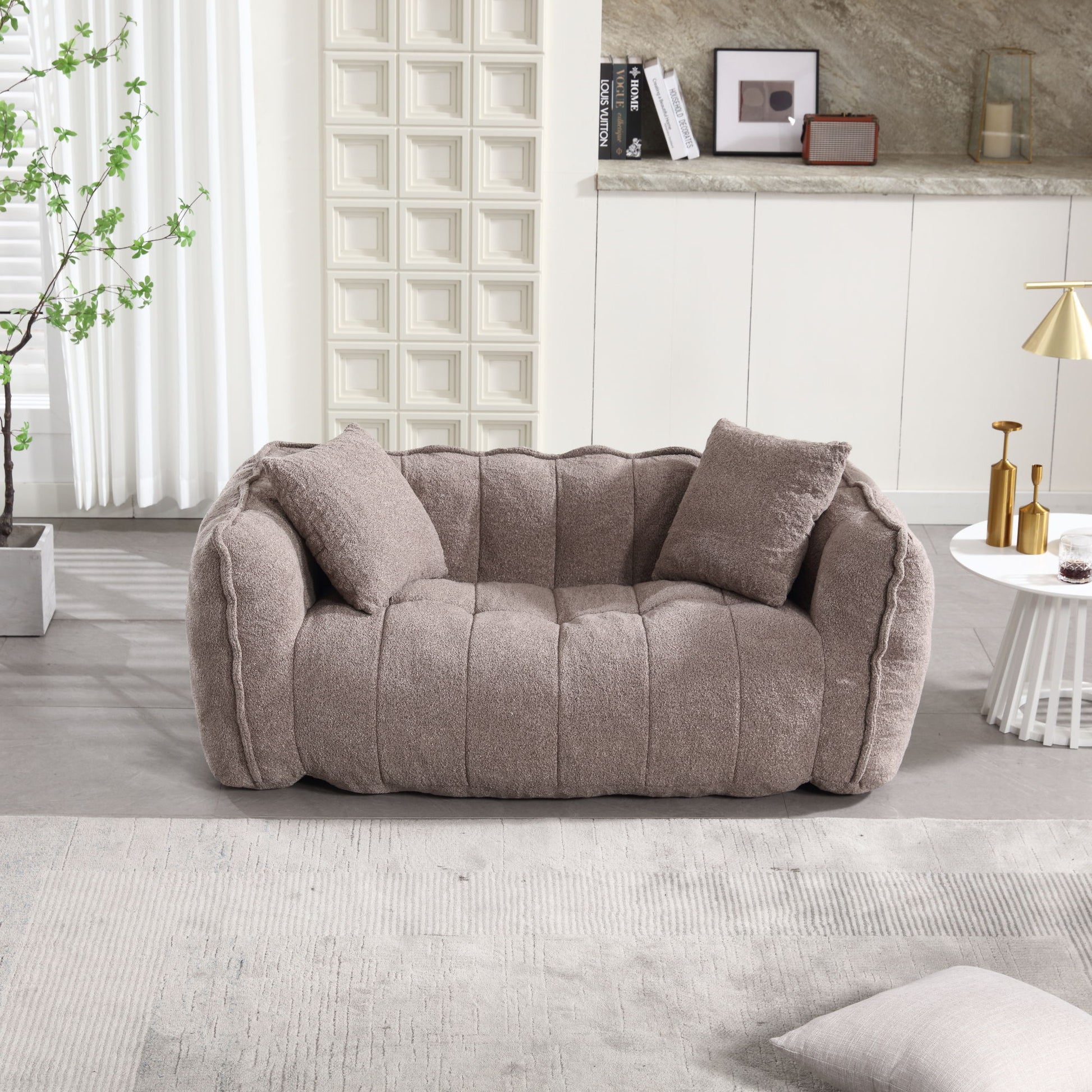 In a modern living room, a soft beanbag chair with high-resilience foam core for two people sits in light brown, accompanied by two matching pillows. A textured wall, decor-enhanced coffee table, plant, and soft lighting define the space. Natural light streams through sheer-curtained windows.