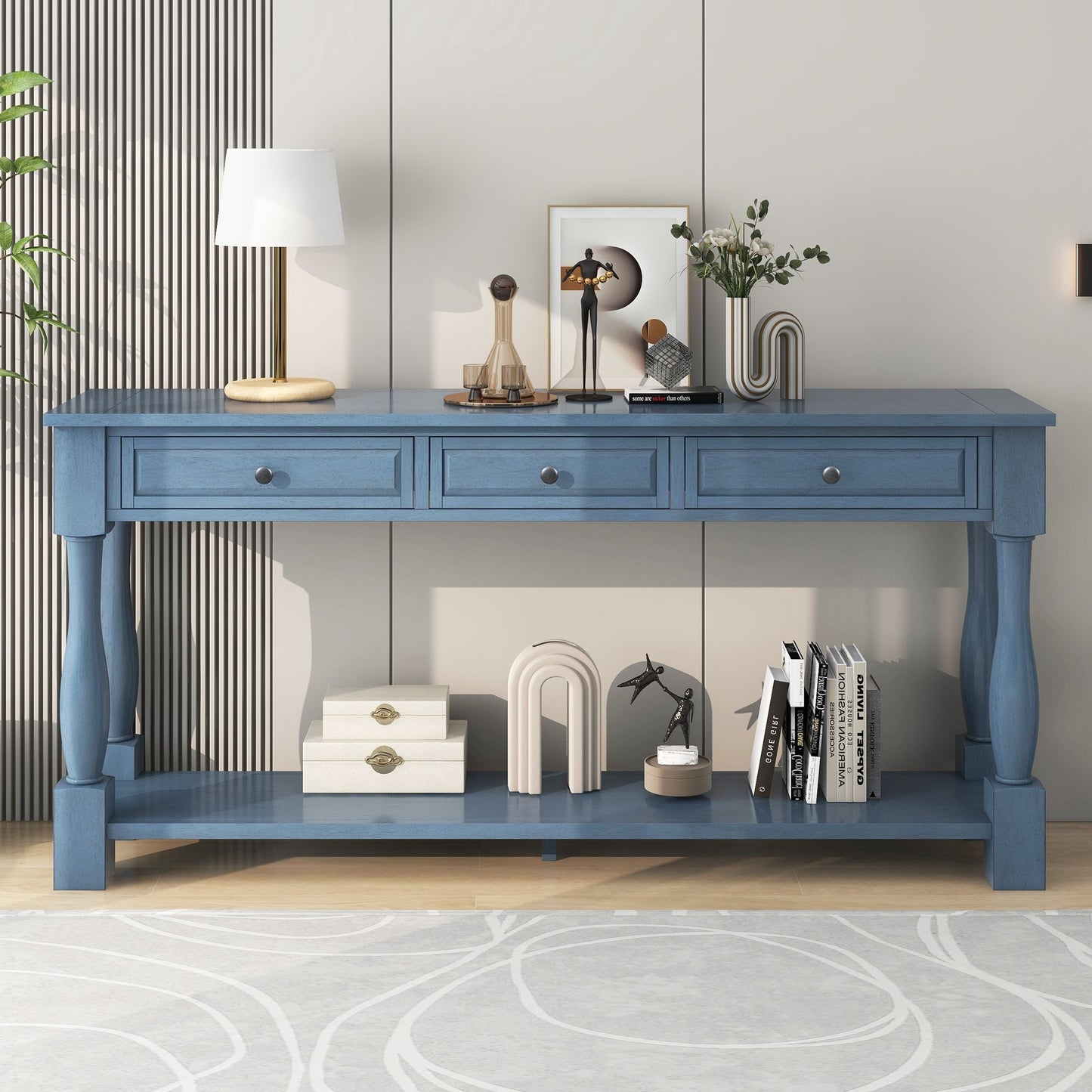 The Long Console Table With Drawers and Shelf stands against a light wall, adorned with a lamp, framed art, flowers, and sculptures. Books and a storage box are neatly arranged below. A circular-patterned rug lies in front.