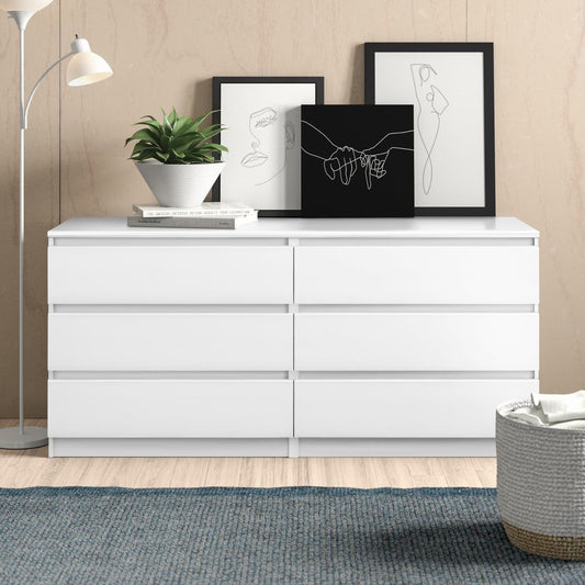 The 6 Drawer Double Dresser for Bedroom Living Room Hallway in white is against a beige wall with three minimalist line art frames, a potted plant, and stacked books. A floor lamp stands to the left on a blue rug over light wood flooring.