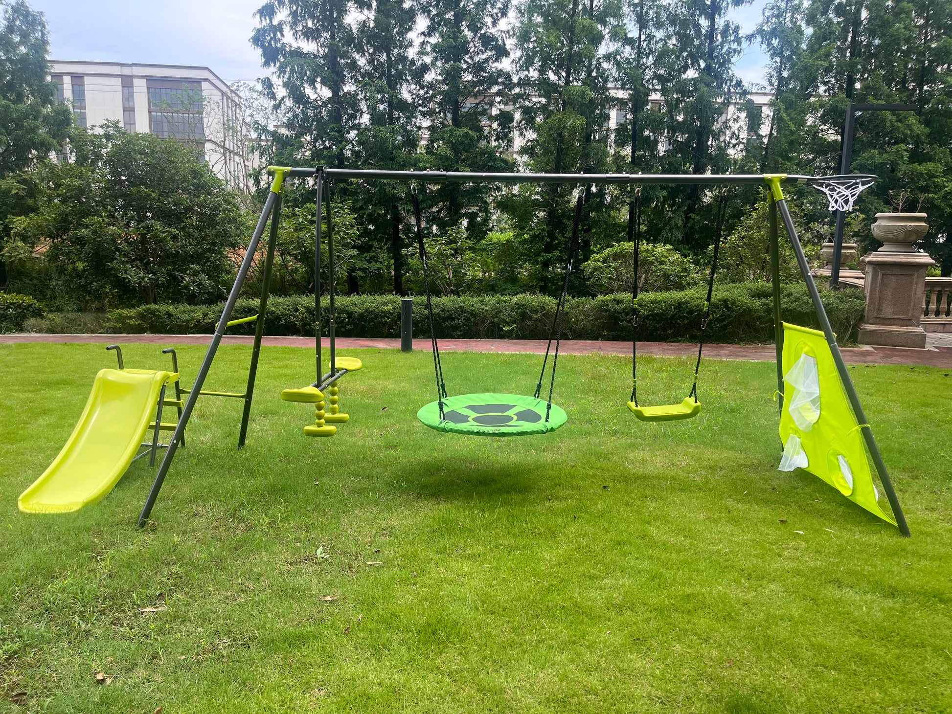 A children's playground set on grass features a yellow slide, teeter-totter, round green swing, two yellow swings, and a basketball hoop. Trees and buildings are visible in the background. Product: Interesting Six Function Swingset - Metal/Plastic, Gray/Blue for ages 3+, 440 lbs capacity with 31.5" net swing.