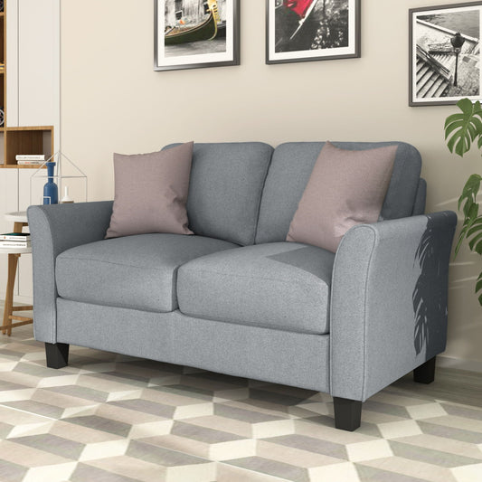 A gray Living Room Furniture Love Seat Sofa with two pink cushions adorns the living room. Three framed pictures decorate the wall behind, while a potted plant is partially visible to the right. The floor showcases a geometric-patterned rug.