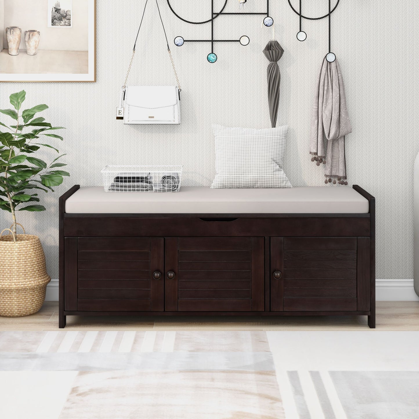 A Storage Bench with 3 shutter-shaped doors and a removable cushion sits against the wall, featuring hidden storage. Above it, decorative hooks hold a white purse and scarf. A potted plant and framed picture add decor, while a light gray rug covers the floor.