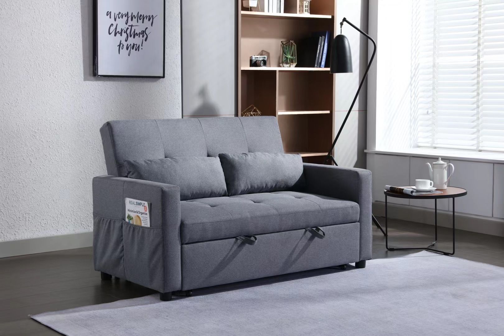 A 2-seater sleeper sofa bed in gray linen fabric, with side pocket storage, is in a living room. Behind it is a wooden shelf with books and decor. Nearby stands a black floor lamp, while a small round table holds a teapot and cup on the rug-covered dark wood floor.