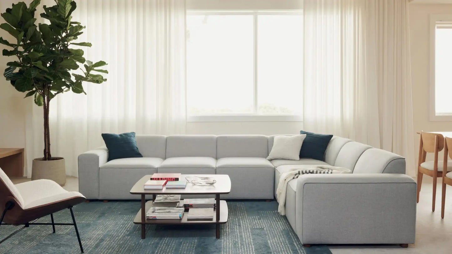 A modern living room featuring the Mambo Loveseat in light gray, adorned with two dark blue cushions, complemented by a wooden coffee table resting on a blue rug. A large window with sheer curtains allows natural light to fill the space. On the left, there's a potted plant, and a wooden chair is partially visible.