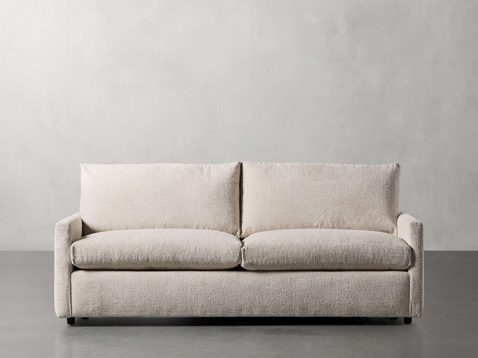 The Kipton 84" Deep Sofa in beige, featuring a minimalist and modern design with plush cushions, clean lines, and armrests, is positioned against a plain gray wall on a smooth concrete floor.