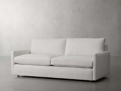 The Kipton 84" Deep Sofa, in a modern white design with clean lines, is set against a plain, light gray background. It boasts a simple aesthetic with two seat cushions and two backrest cushions, delivering a minimalist and contemporary appearance.