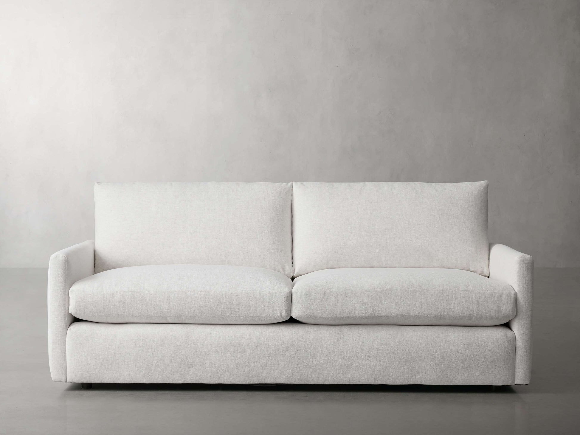 The Kipton 84" Deep Sofa, featuring a modern, minimalist design with clean lines and upholstered in light gray fabric, is set against a plain, light gray wall. It includes two large seat cushions and two back cushions, offering a simple and elegant appearance.