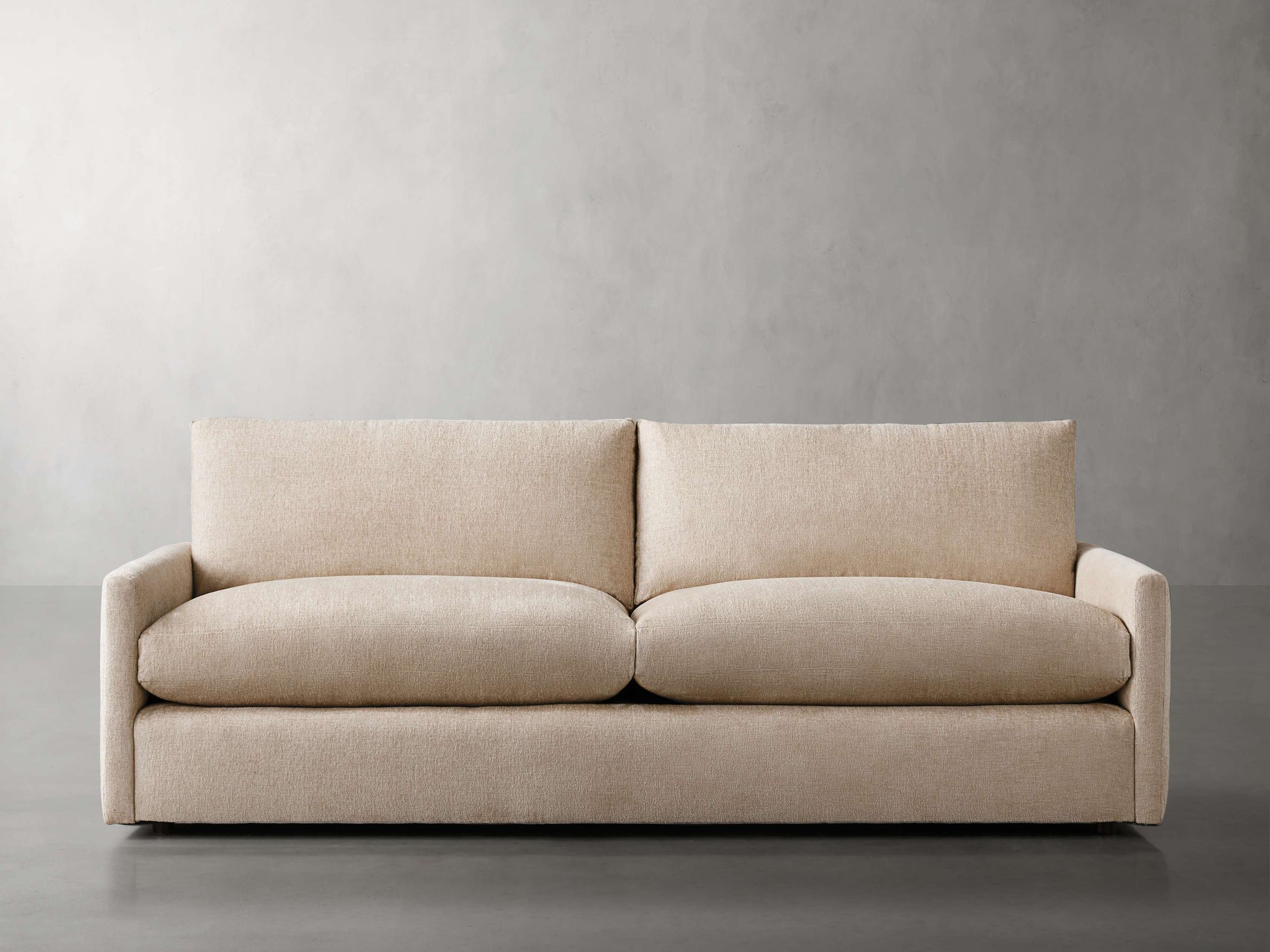 The Kipton 84" Deep Sofa, with its minimalist beige design and plush cushions, is positioned against a plain gray backdrop. Boasting a modern aesthetic with straight lines and soft fabric upholstery, it exudes a simple yet elegant look.