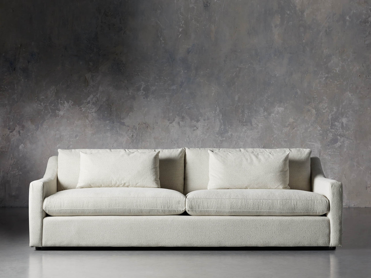 The Ashby Sofa, a minimalist off-white design featuring two large cushions, sits against a textured gray wall with concrete flooring.