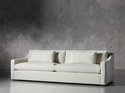 The Ashby Sofa, featuring a modern design with two large seat cushions and two back cushions in a light color, is set against a textured gray concrete wall on a smooth, light gray floor. Its minimalist style exudes an elegant and contemporary flair.