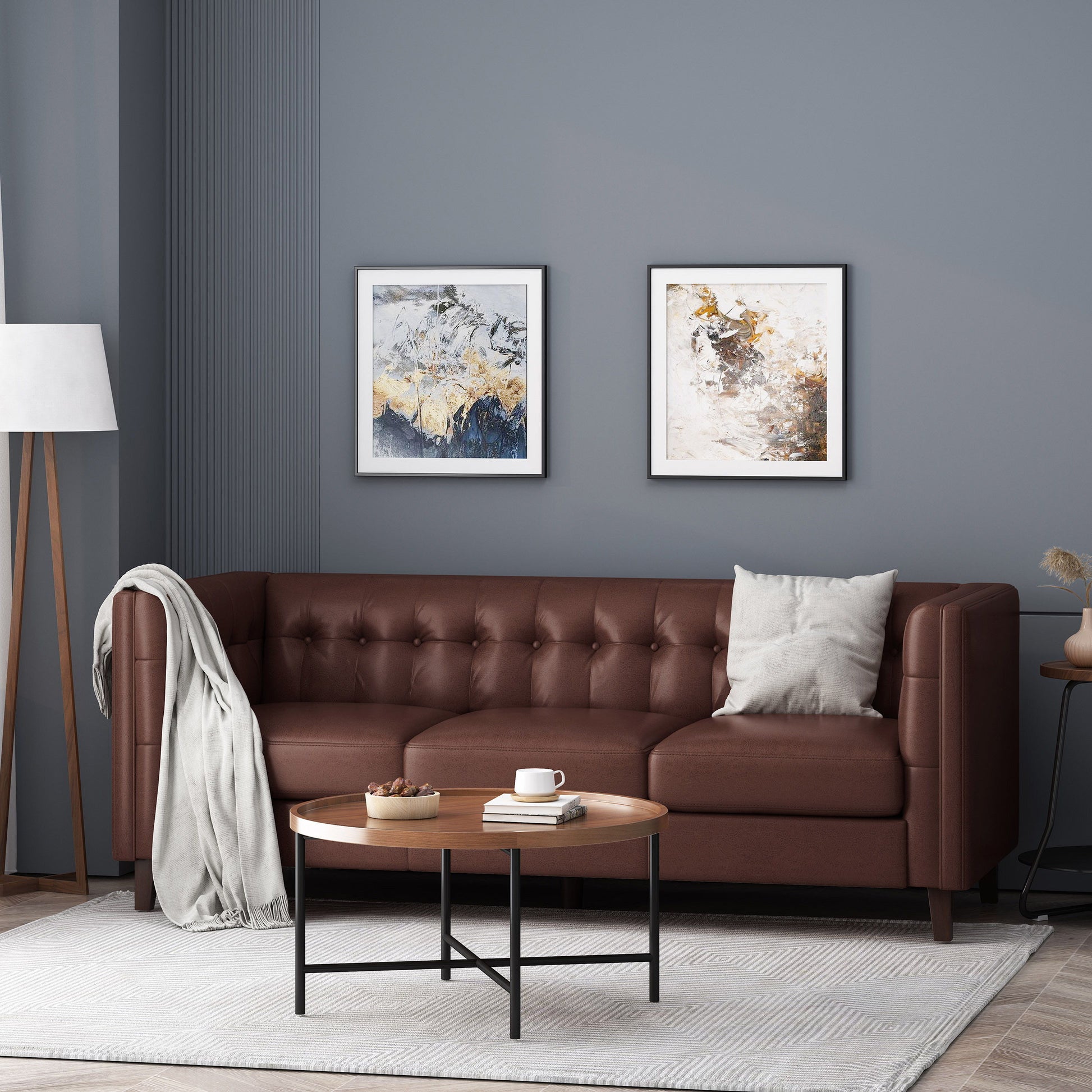 A modern living room features the Comfy 3 Seat Sofa with tufted back in brown leather, a gray throw, and a beige cushion. Two abstract paintings adorn the gray wall above. A round wooden coffee table sits on a white textured rug, with decor. A floor lamp brightens the corner.