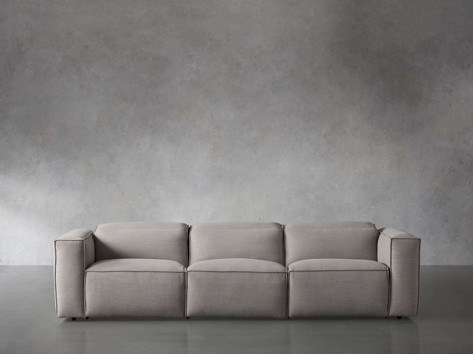 The Coburn Three Piece Motion Sofa, featuring a sleek modern design and minimalist gray tones, is positioned in front of a textured gray concrete wall. The room exudes an industrial aesthetic complemented by the smooth gray flooring.