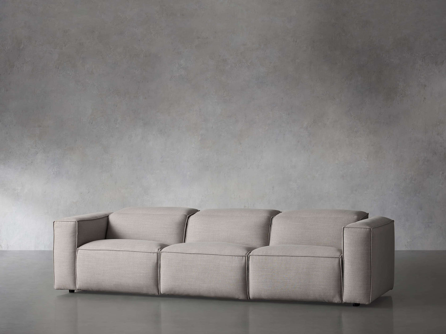 The Coburn Three Piece Motion Sofa, with its modern and minimalist design, features three seat cushions upholstered in gray fabric. It seamlessly complements the textured gray walls and floor, offering a sleek and simple aesthetic that cultivates a calm and neutral atmosphere in the space.