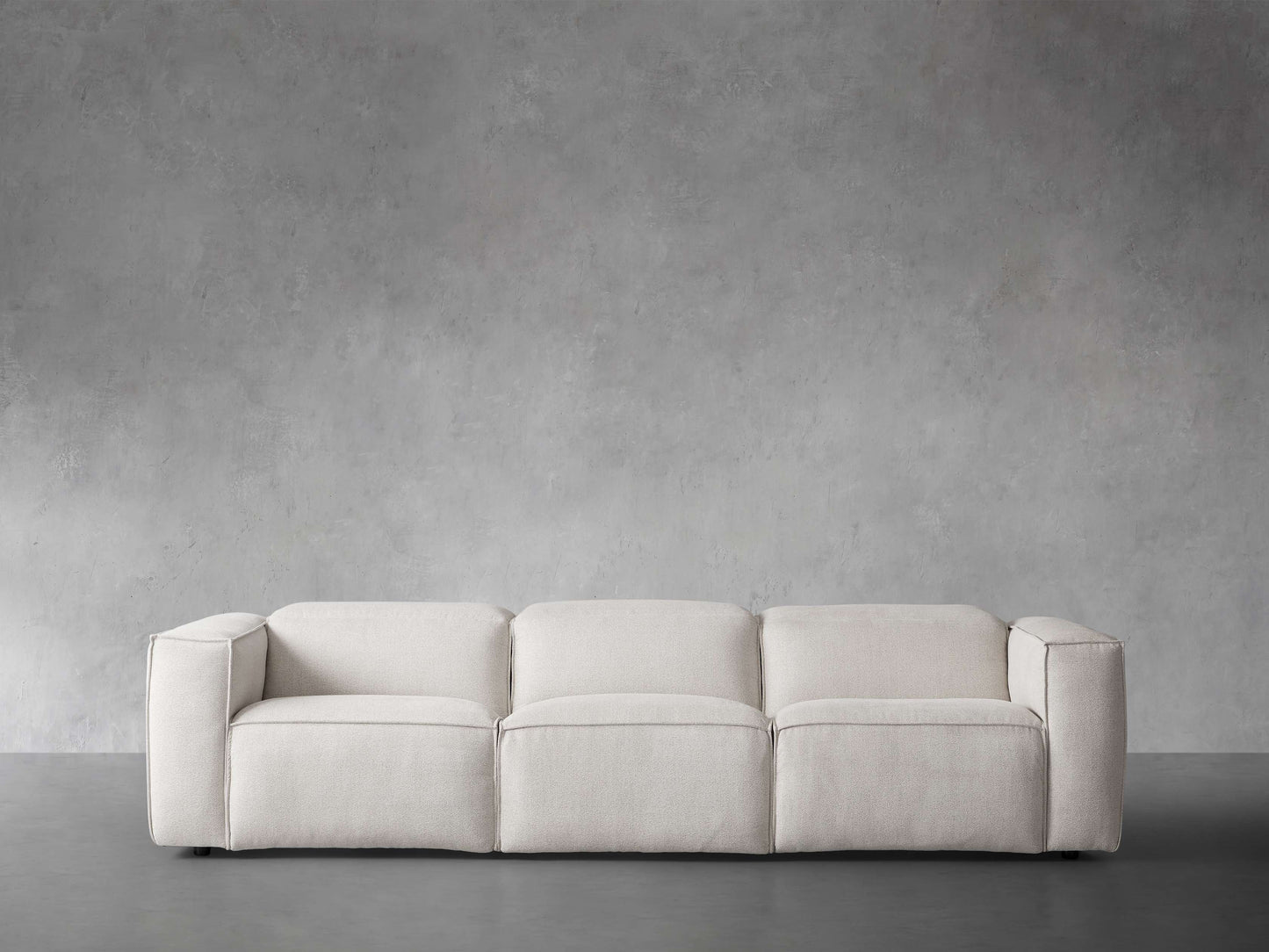 The Coburn Three Piece Motion Sofa, in a modern and minimalist beige design, is elegantly set against a plain gray concrete wall. Its clean lines and low profile contribute to a contemporary and inviting atmosphere.