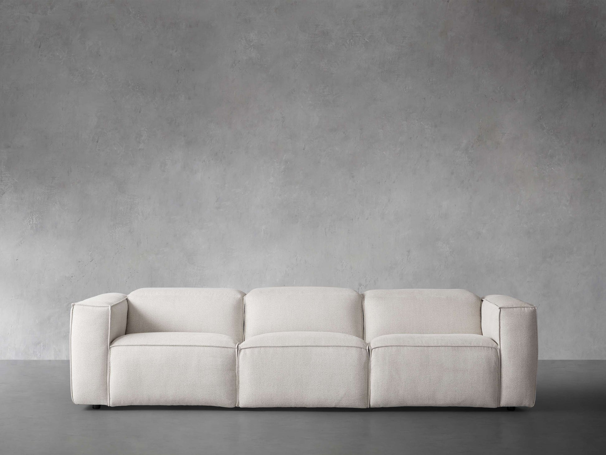 The Coburn Three Piece Motion Sofa, in a modern and minimalist beige design, is elegantly set against a plain gray concrete wall. Its clean lines and low profile contribute to a contemporary and inviting atmosphere.