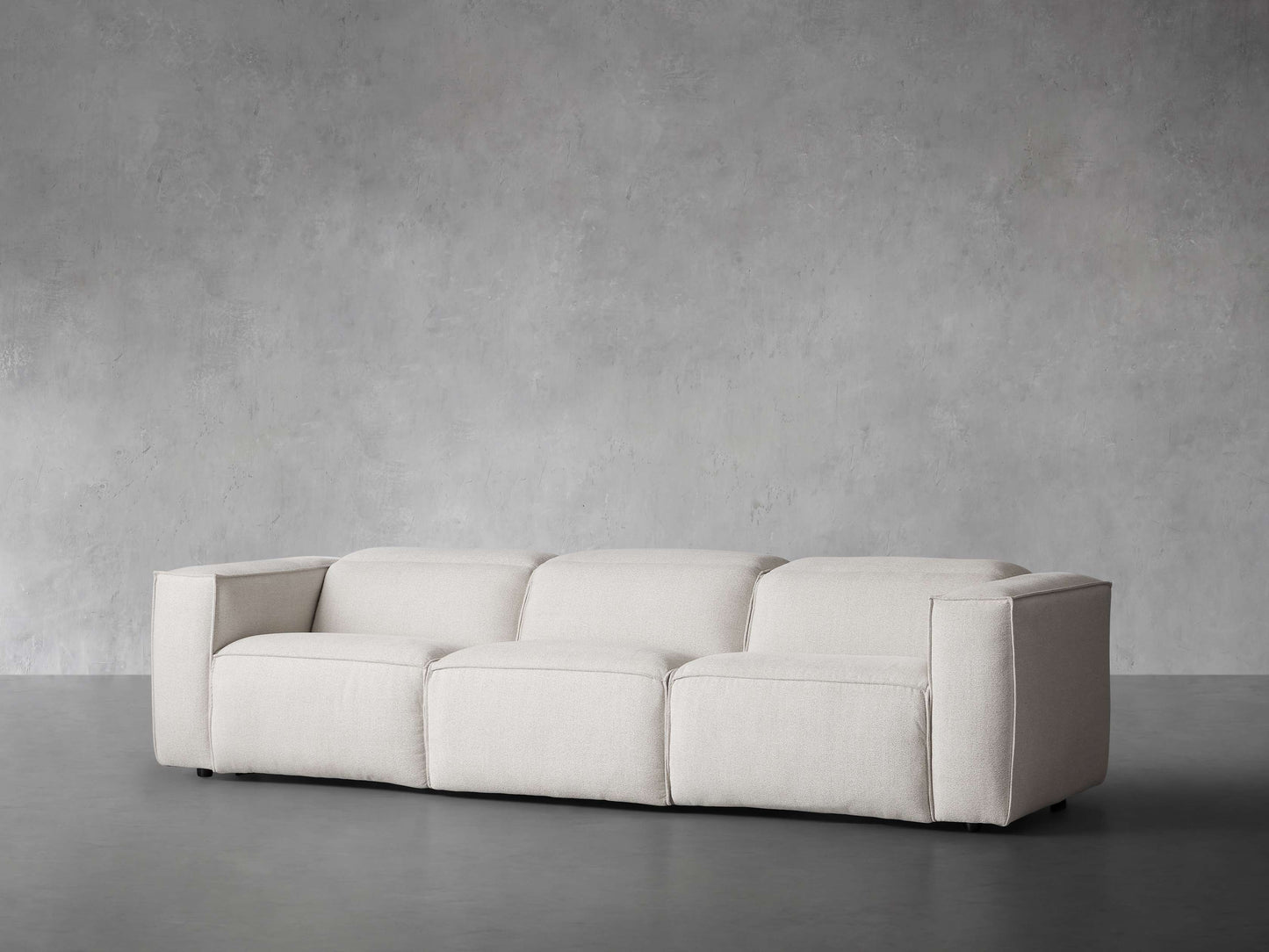 The Coburn Three Piece Motion Sofa, with its minimalist beige design and clean lines, is set against a plain gray concrete wall. The sofa's wide armrests and modern aesthetic contribute to a sleek and contemporary look in the room.