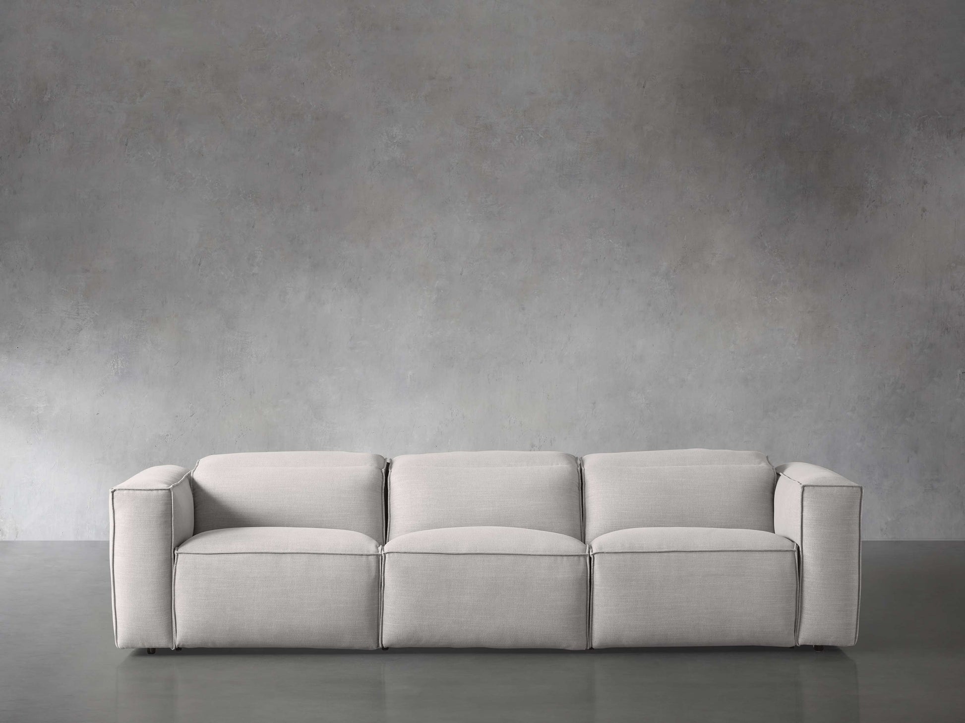 The Coburn Three Piece Motion Sofa, with its modern and minimalist design and accompanied by three cushions, is placed against a textured grey wall on a smooth grey floor, creating a seamless and monochromatic look.