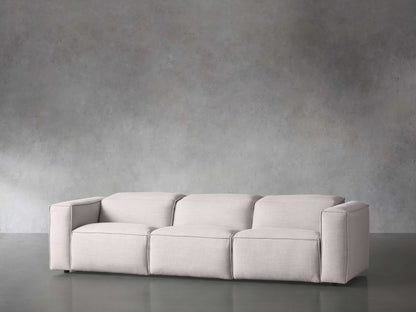 The Coburn Three Piece Motion Sofa in a sleek light gray hue is positioned against a textured gray wall. The room showcases a minimalist design accentuated by a smooth, reflective gray floor.