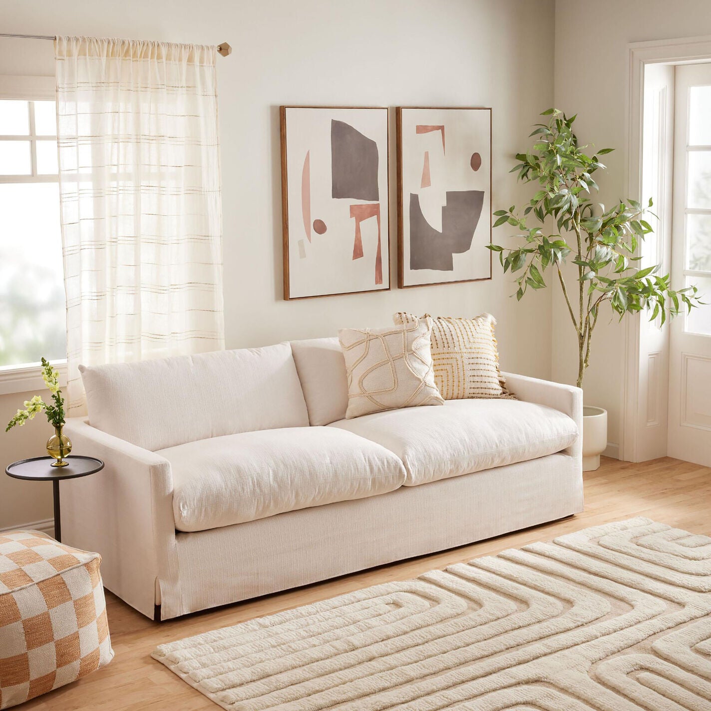 A cozy living room featuring the Brynn Feather-Filled Sofa, dressed with patterned cushions, sits atop a light-colored rug displaying geometric designs. The walls are adorned with abstract paintings, complemented by a tall potted plant and a small side table holding a vase.