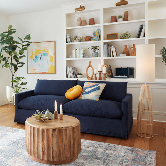 A cozy living room featuring the Brynn Feather-Filled Sofa in navy blue, adorned with colorful cushions, a round wooden coffee table, and a tall floor lamp. The background showcases white built-in shelves filled with books, decorative items, and a potted plant, while a vibrant abstract painting hangs on the wall.