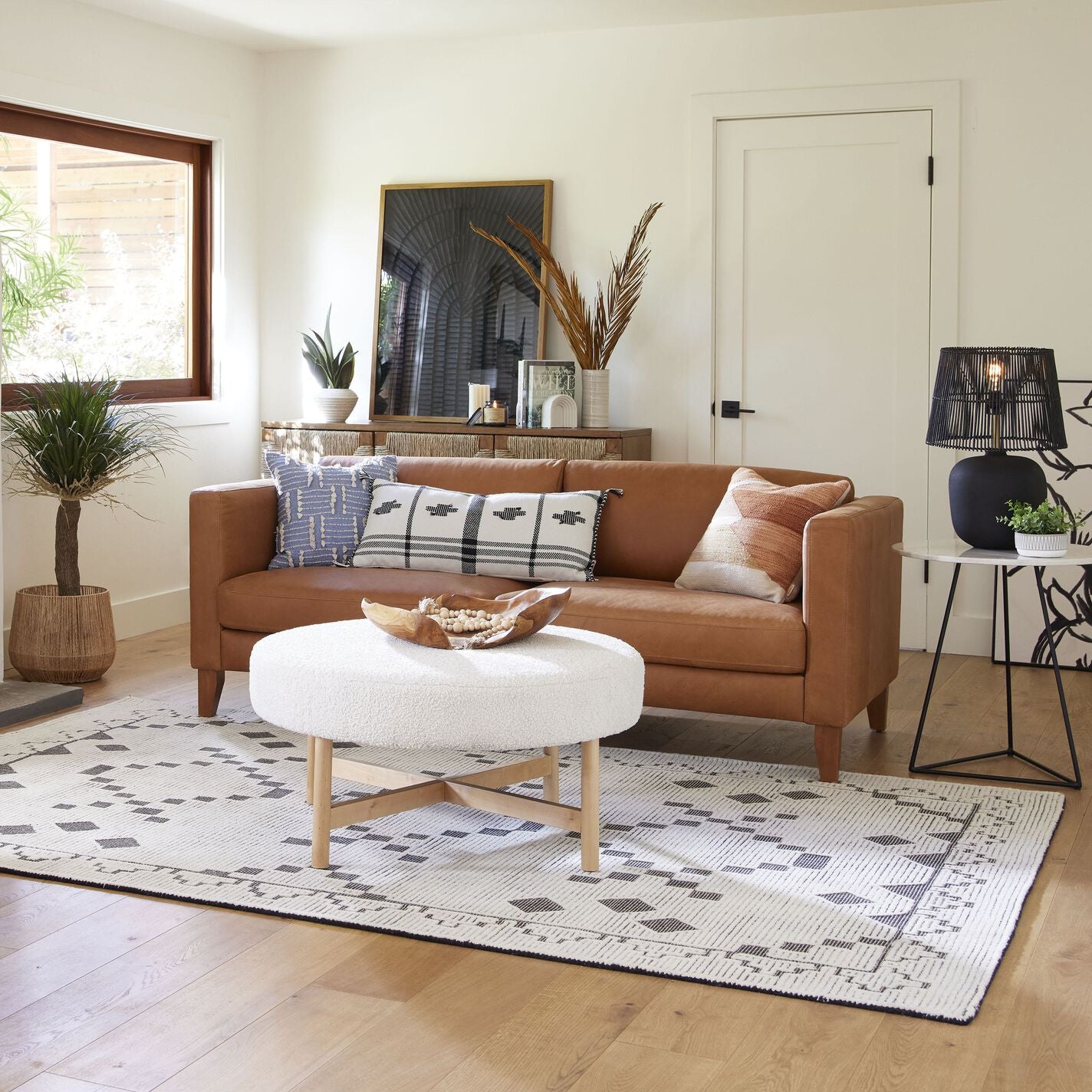 A cozy living room featuring the Abrie Vintage Tan Leather Sofa adorned with various patterned pillows and a round white coffee table. The space is decorated with plants, a patterned rug, and a framed artwork on the shelf. Natural light filters in through a large window.