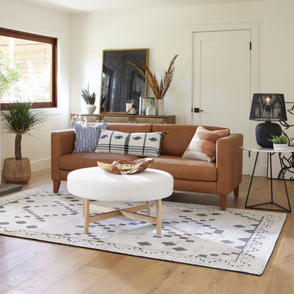 A cozy living room featuring the Abrie Vintage Tan Leather Sofa adorned with various patterned pillows and a round white coffee table. The space is decorated with plants, a patterned rug, and a framed artwork on the shelf. Natural light filters in through a large window.