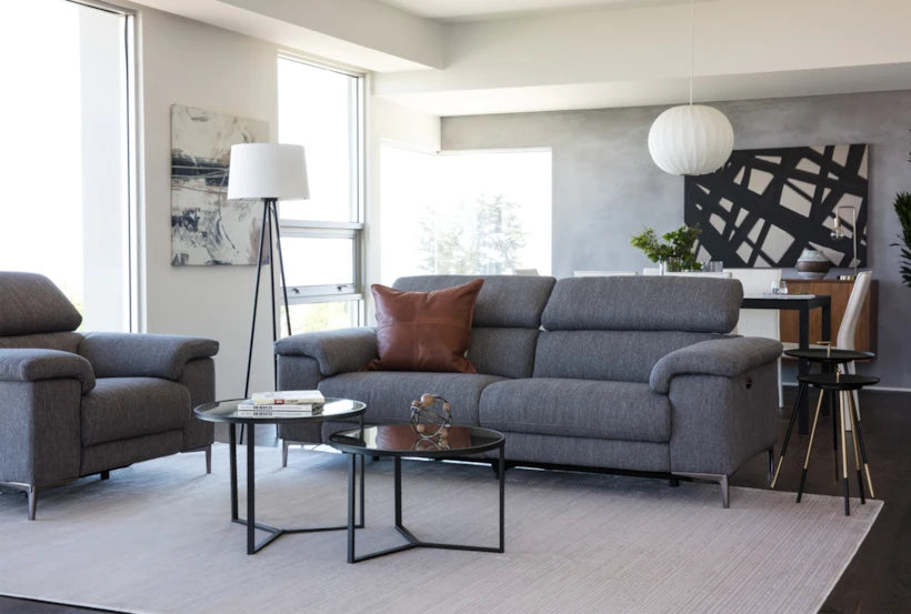 An effortlessly stylish living room featuring the Talin Power Reclining Sofa with USB in grey, complemented by a brown cushion and illuminated by a sleek white floor lamp on a light rug. Two round glass coffee tables display decorative pieces while the dining area in the background is anchored by a sophisticated dining table with chairs and an elegant pendant light. Abstract art adorns the walls, and natural light flows in beautifully.