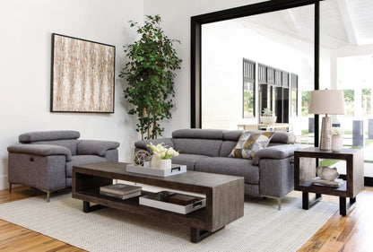 A modern living room features the Talin Power Reclining Sofa with USB in gray alongside an armchair, complemented by a wooden coffee table and a potted plant. A large sliding glass door offers a glimpse into an outdoor area. A framed abstract art piece adorns the wall, while a table lamp rests on a nearby side table.