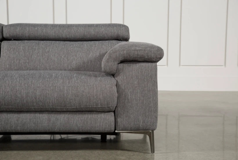 The Talin Power Reclining Sofa with USB, featuring a modern grey fabric design, adjustable headrest, metal legs, and an armrest, is set against a white paneled wall and a polished concrete floor.