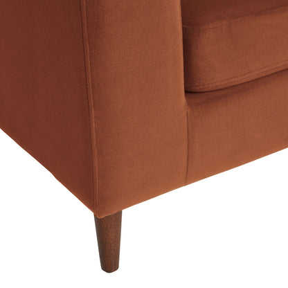 A close-up view of a corner of the rust-colored Camile Velvet Right-Facing Sectional Sofa showcases the upholstery texture and the wooden leg supporting the sofa. The design exhibits clean, simple lines with a minimalist aesthetic.