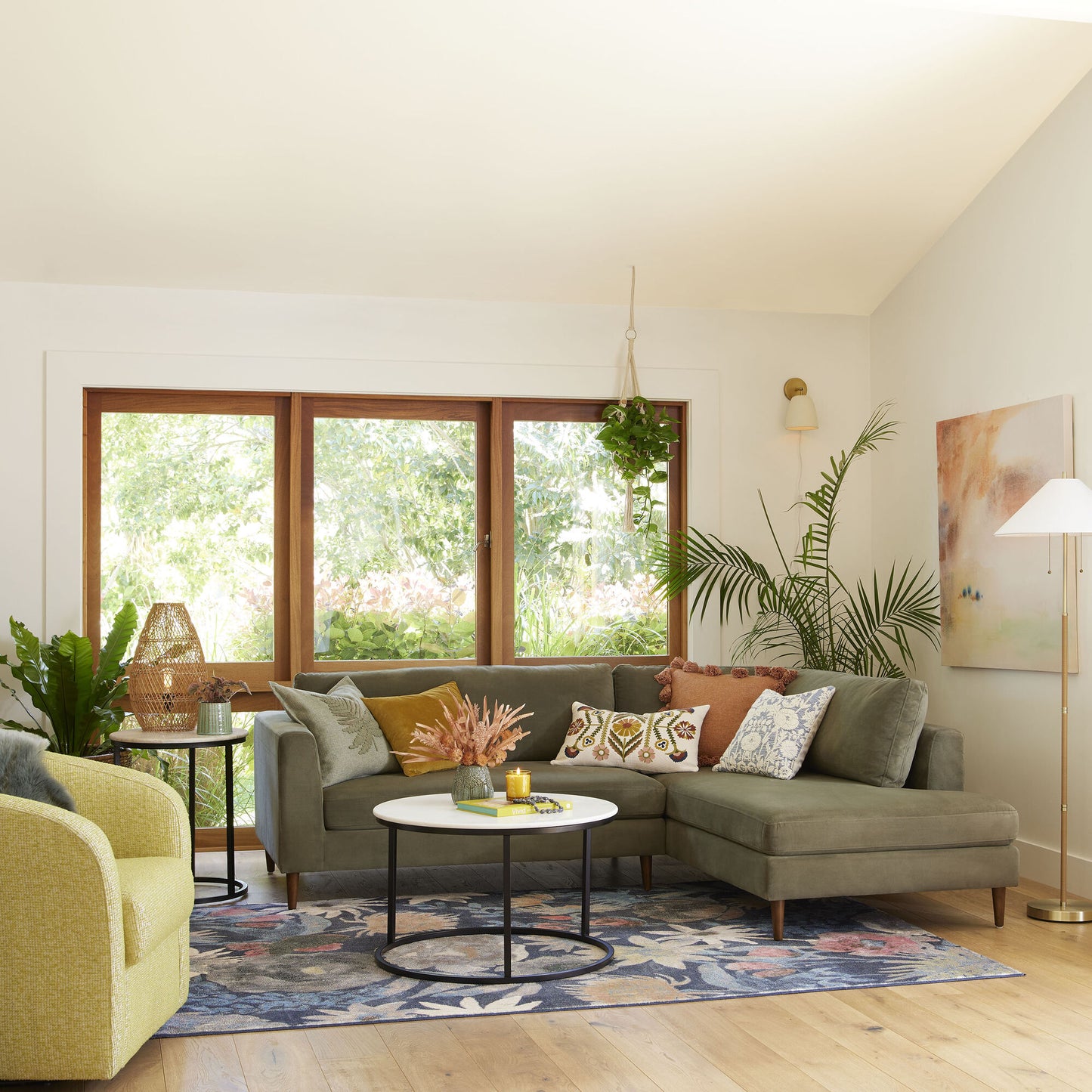 A bright living room featuring a Camile Velvet Right-Facing Sectional Sofa, adorned with colorful pillows and a floral rug. The space includes plants, a round coffee table, a floor lamp, and a painting. Large windows invite natural light in, while a hanging plant enhances the room's natural vibe.