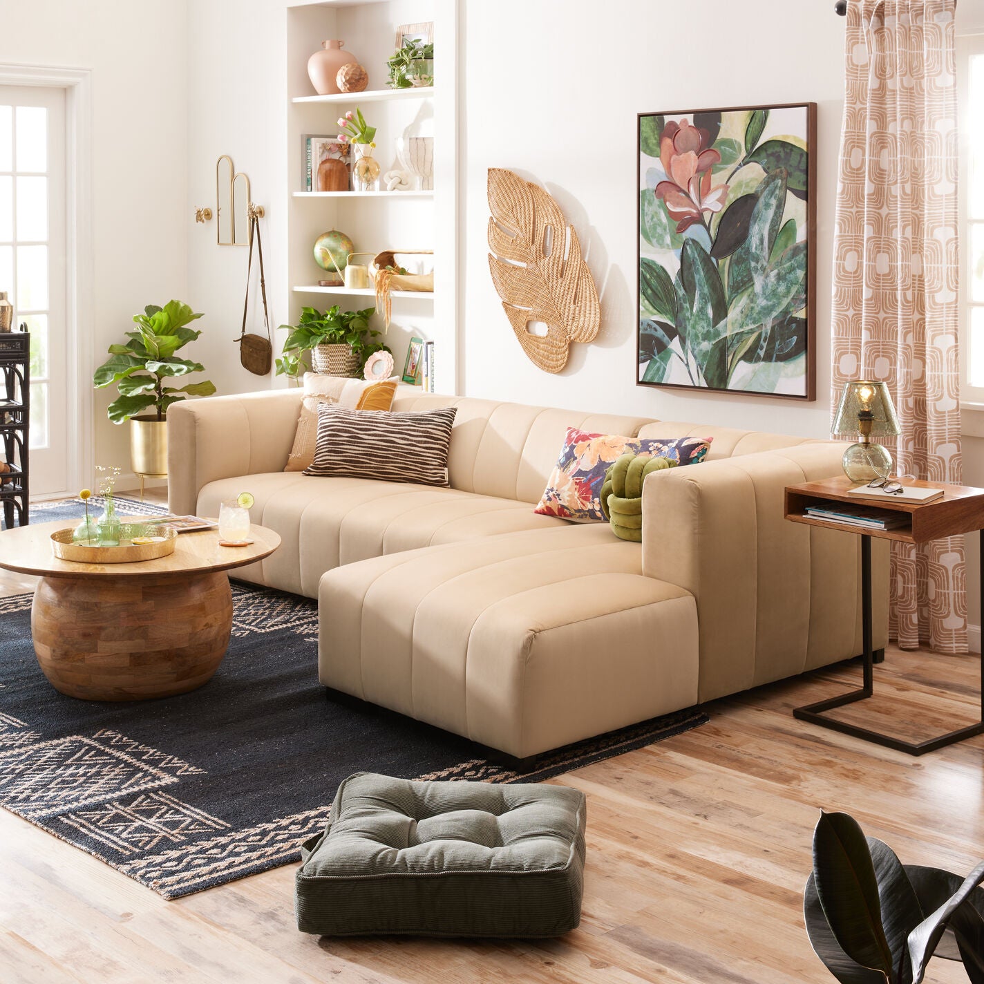 A cozy living room featuring a spacious Cambrie Wheat Velvet Right-Facing Sectional Sofa, a round wooden coffee table, and tropical-themed decor. Shelves display plants and vases, while a botanical artwork hangs on the wall. A patterned rug covers the wood floor.