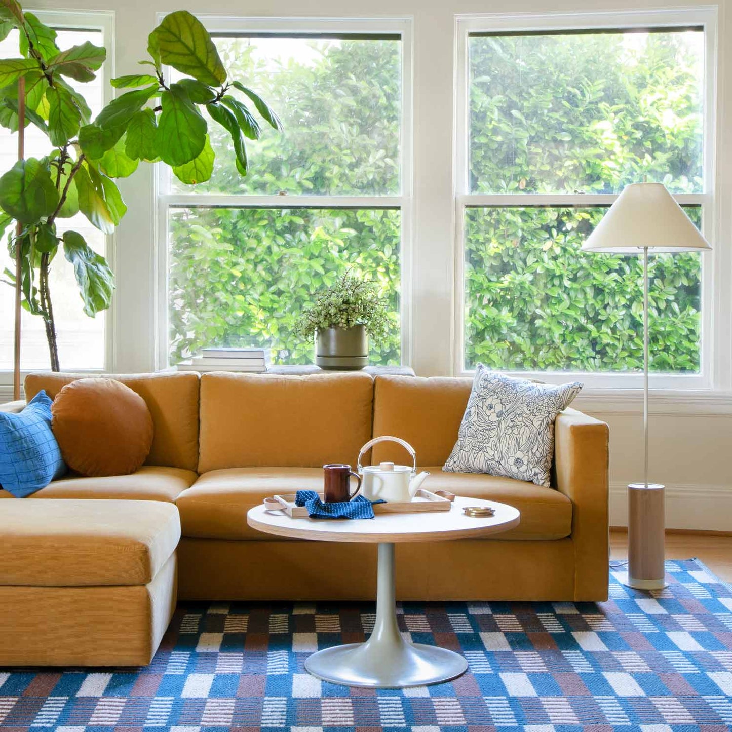 A cozy living room featuring the Milo Sofa in a rich mustard yellow, embellished with blue and patterned pillows. A white coffee table supports a teapot and mugs. Large windows offer views of lush greenery, while a tall floor lamp is positioned beside the sofa on a blue-patterned rug.