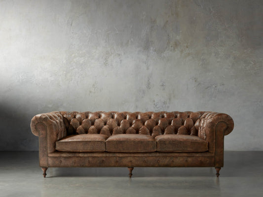 A Wessex Leather Sofa, featuring tufted brown leather and rolled arms, is positioned against a textured gray wall on a polished concrete floor. With its three cushions and wooden legs, it exudes a classic and elegant appearance.