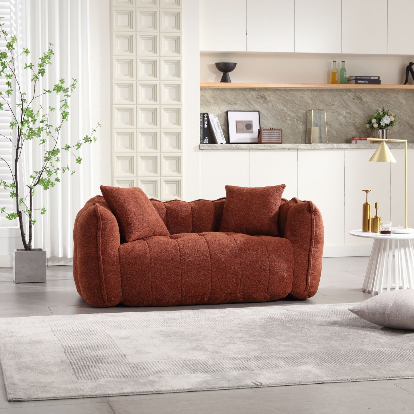 A cozy, soft beanbag chair with high-resilience foam core for two is set in a modern living room. The backdrop includes white cabinets, a light marble backsplash, decor on a shelf, and a potted plant. Nearby is a floor lamp and small table on a light gray rug.