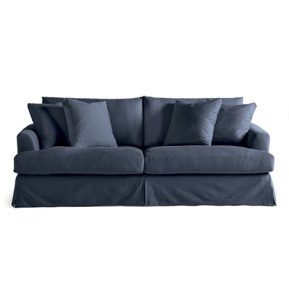 Introducing the Ava Slipcovered Sofa, a dark blue, two-seat sofa complemented by multiple matching cushions. This sofa showcases a skirted base with a plush and comfortable appearance, beautifully set against a white background.