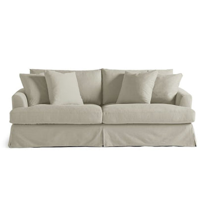 The Ava Slipcovered Sofa, featuring a light beige upholstery with a skirted base, is displayed. It includes three seat cushions and five matching throw pillows, offering a cozy and comfortable look. The sofa is set against a plain white background.