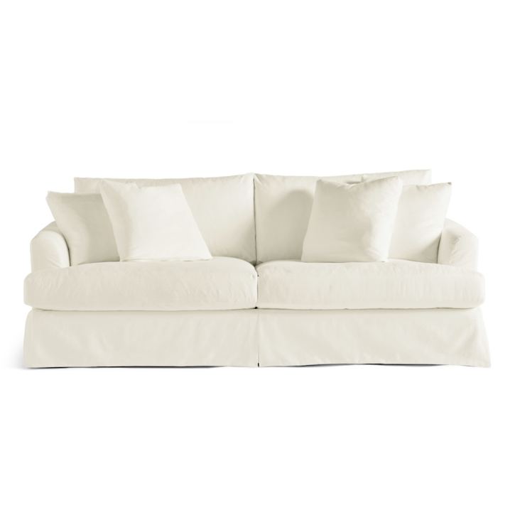 The Ava Slipcovered Sofa is cream-colored and comes with plush cushions and a skirted base. It has a comfortable, overstuffed appearance, featuring two main seat cushions and several matching throw pillows against a plain white backdrop.