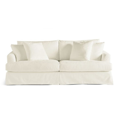 The Ava Slipcovered Sofa is cream-colored and comes with plush cushions and a skirted base. It has a comfortable, overstuffed appearance, featuring two main seat cushions and several matching throw pillows against a plain white backdrop.