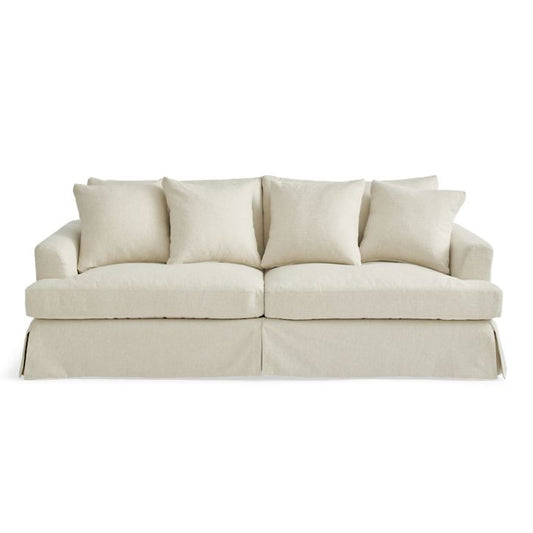 The Ava Slipcovered Sofa is a beige masterpiece accompanied by four matching cushions, showcasing a skirted design at the bottom. Both the cushions and sofa exude a minimalist and modern aesthetic, offering a cozy yet elegant appearance.