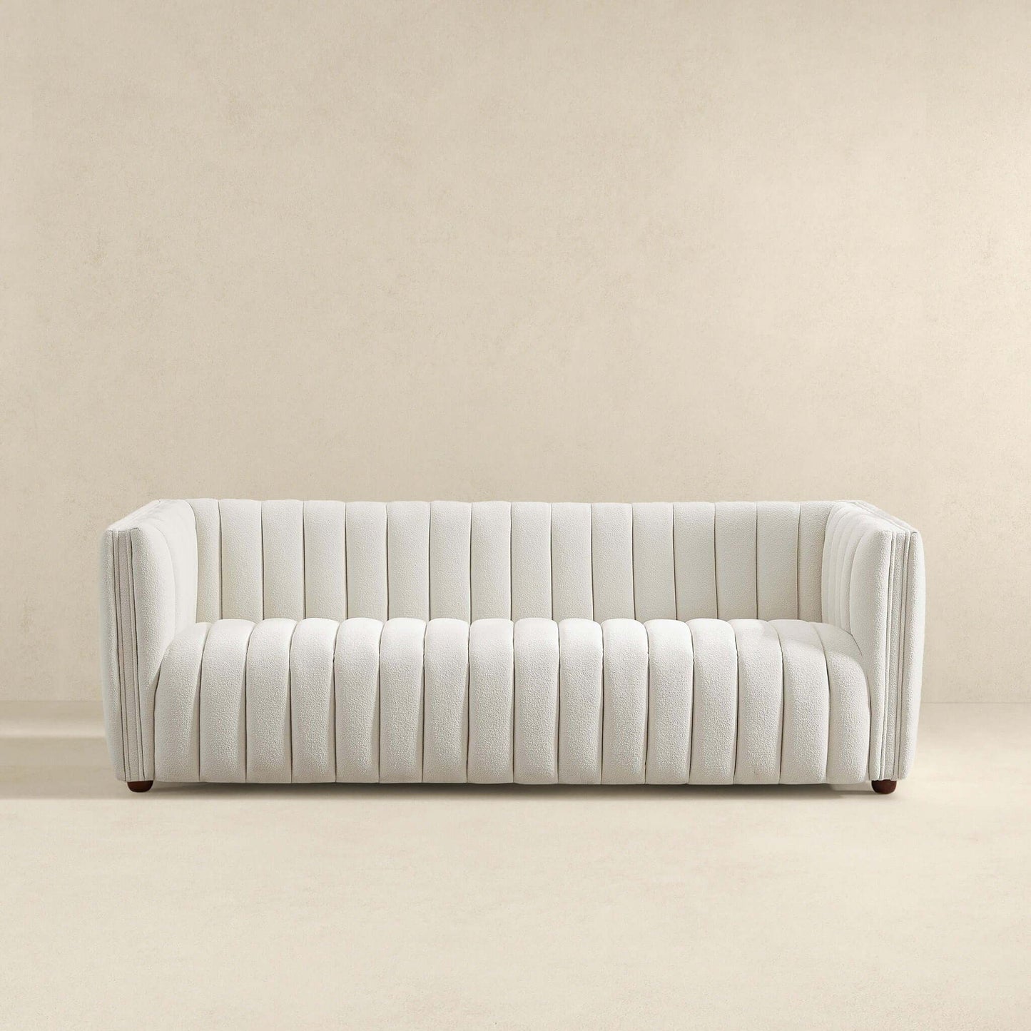 A minimalist white April - Mid-Century Modern Luxury Tight Back Boucle Couch with a ribbed, tufted design sits against a plain beige wall. It boasts a contemporary low-profile look with square arms and small, barely visible round legs.