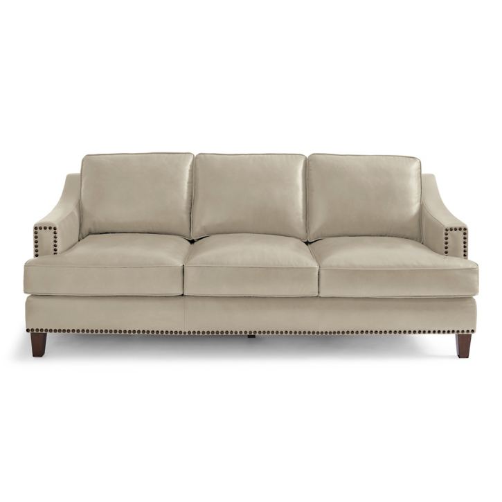 The Collins Sofa is a beige three-seater featuring plush cushions, nailhead trim on the arms and base, and tapered wooden legs.